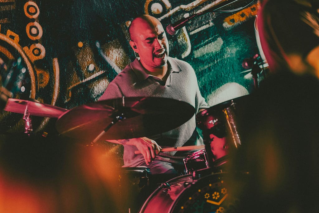 Fotografía profesional de la banda de rock Los Martes en concierto en KUP, Centro Histórico de Mexicali, Baja California. Imagen con estilo editorial y estética rockera grunge, capturada por el fotógrafo Yasunari Murayama con cámara Canon R6. Escenario decorado con arte urbano, luces de concierto, público disfrutando la música en vivo. Ideal para portadas de revista musical y documental de eventos. Fotografía artística de banda independiente en Mexicali, foto de guitarrista en escena, ambiente nocturno, rock alternativo, cultura musical local, músicos tocando en vivo, show energético, fotografía para promoción de bandas, imagen para cartel de concierto, retrato de rock en acción.