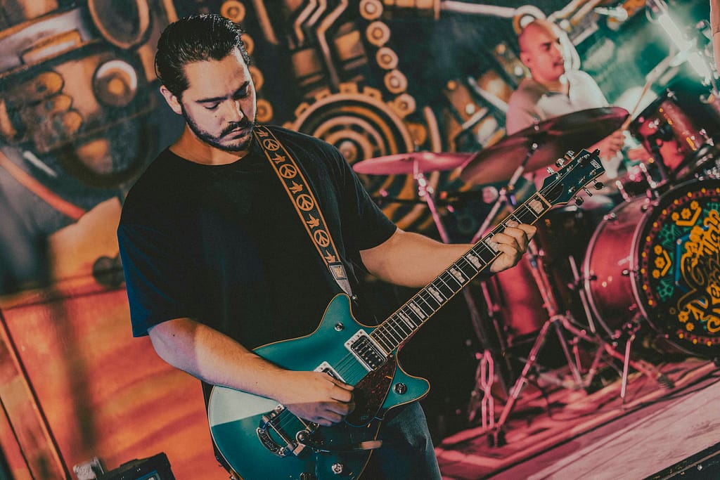 Fotografía profesional de la banda de rock Los Martes en concierto en KUP, Centro Histórico de Mexicali, Baja California. Imagen con estilo editorial y estética rockera grunge, capturada por el fotógrafo Yasunari Murayama con cámara Canon R6. Escenario decorado con arte urbano, luces de concierto, público disfrutando la música en vivo. Ideal para portadas de revista musical y documental de eventos. Fotografía artística de banda independiente en Mexicali, foto de guitarrista en escena, ambiente nocturno, rock alternativo, cultura musical local, músicos tocando en vivo, show energético, fotografía para promoción de bandas, imagen para cartel de concierto, retrato de rock en acción. productor de video en mexicali yasunari murayama