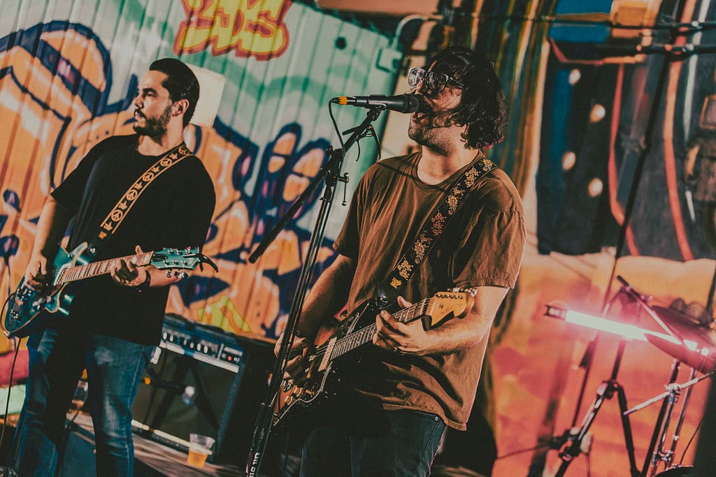 Concierto de Los Martes en vivo en KUP, Mexicali, Baja California. Foto de guitarrista tocando con energía, estilo grunge y estética de revista rockera. Fotografía profesional capturada por Yasunari Murayama con Canon R6, luces cálidas y fondo de mural artístico. Imagen ideal para medios musicales y redes sociales de la banda. Documental de música en vivo, evento cultural en el centro histórico de Mexicali. Rock alternativo, escena independiente, fotografía de espectáculo en vivo, retrato de músico en concierto, ambiente bohemio, estilo visual cinematográfico, fotografía editorial para promoción musical, foto para prensa cultural y agenda de eventos. productor de video en mexicali yasunari murayama