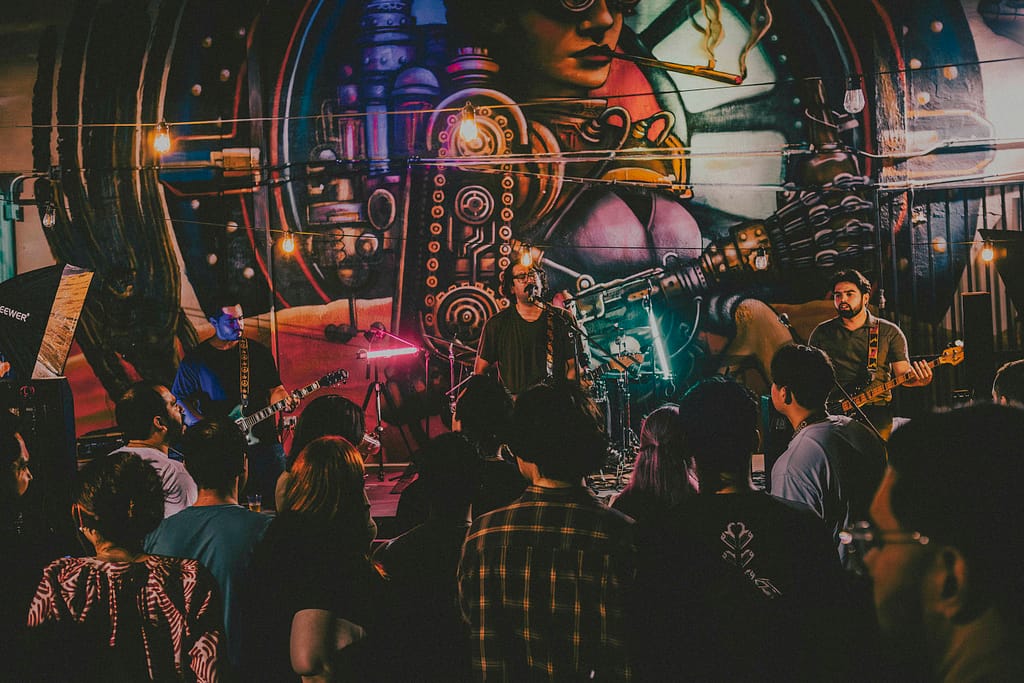 Fotografía profesional de la banda de rock Los Martes en concierto en KUP, Centro Histórico de Mexicali, Baja California. Imagen con estilo editorial y estética rockera grunge, capturada por el fotógrafo Yasunari Murayama con cámara Canon R6. Escenario decorado con arte urbano, luces de concierto, público disfrutando la música en vivo. Ideal para portadas de revista musical y documental de eventos. Fotografía artística de banda independiente en Mexicali, foto de guitarrista en escena, ambiente nocturno, rock alternativo, cultura musical local, músicos tocando en vivo, show energético, fotografía para promoción de bandas, imagen para cartel de concierto, retrato de rock en acción. productor de video en mexicali yasunari murayama