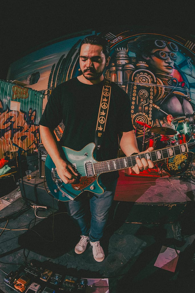 Concierto de Los Martes en vivo en KUP, Mexicali, Baja California. Foto de guitarrista tocando con energía, estilo grunge y estética de revista rockera. Fotografía profesional capturada por Yasunari Murayama con Canon R6, luces cálidas y fondo de mural artístico. Imagen ideal para medios musicales y redes sociales de la banda. Documental de música en vivo, evento cultural en el centro histórico de Mexicali. Rock alternativo, escena independiente, fotografía de espectáculo en vivo, retrato de músico en concierto, ambiente bohemio, estilo visual cinematográfico, fotografía editorial para promoción musical, foto para prensa cultural y agenda de eventos.